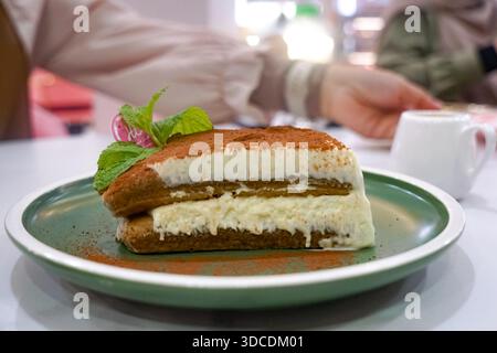 Ricca fetta di tiramisù con strati di spugna imbevuti di caffè, crema di mascarpone, spolveratura di cacao, foglie di menta e copertura decorativa su piatto verde. Foto Stock