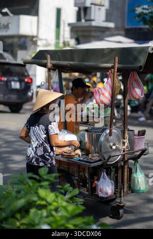 Ho chi Minh City, Vietnam- novembre 2019: Il venditore di strada vietnamita vende cracker di riso in bicicletta, indossando un cappello conico, mostrando autentica vita quotidiana Foto Stock