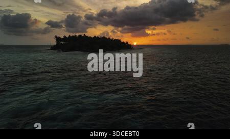 Vista aerea di una silhouette di un'isola tropicale circondata da acque oceaniche scure e ondulate sotto un cielo infuocato al tramonto, Maldive. Foto Stock