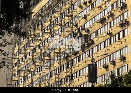 La vista della luce del sole splende dalle finestre ripetitive e dalle unità di aria condizionata di un torreggiante edificio giallo, incorniciato dal fogliame scuro, Chongqing, CH Foto Stock