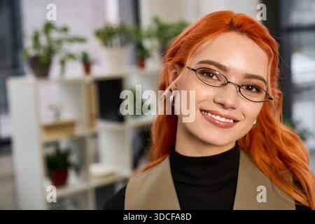La giovane donna con i capelli rossi sorride calorosamente mentre lavora in un ufficio moderno pieno di piante. Foto Stock