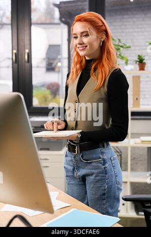 Una donna dai capelli rossi sorride mentre lavora a un progetto in un ambiente di ufficio invitante. Foto Stock