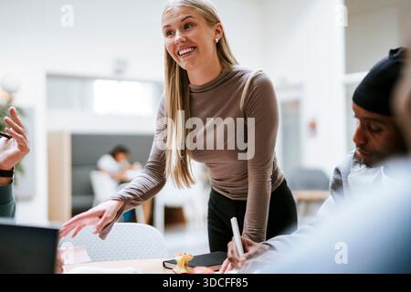Giovane donna dai capelli biondi sorridente che spiega idee di progetto agli amici del campus universitario Foto Stock