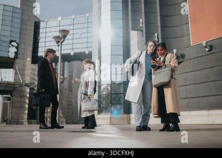 Gli uomini d'affari si incontrano all'esterno di un moderno edificio per uffici, discutendo di progetti con borse e cappotti in un ambiente urbano Foto Stock