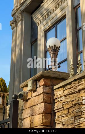 Luce esterna in bronzo ornata con diffusore bianco testurizzato su piedistallo in muratura contro pareti in stucco con pilastri e telecamera protettiva. Foto Stock
