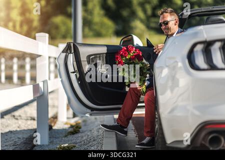 Un uomo siede su un'auto bianca con un mazzo di rose, preparandosi a uscire in una giornata di sole a bordo strada. Foto Stock