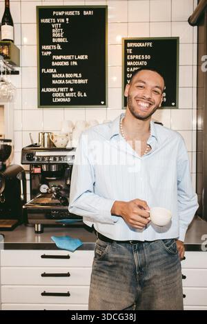 Ritratto del barista sorridente che tiene la tazza di caffè in piedi vicino al bancone del bar Foto Stock