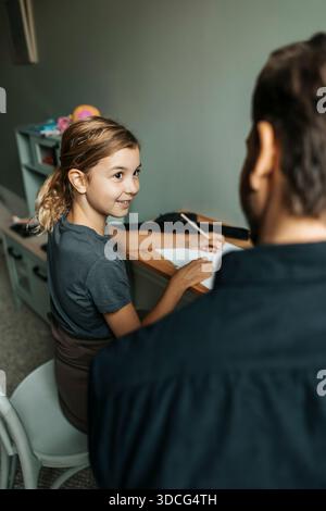 Ragazza sorridente che fa i compiti con l'aiuto del padre a casa Foto Stock