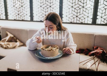 Una donna in un comodo maglione bianco gode di una ciotola di pasta in un ambiente rilassato Foto Stock