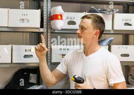Uomo in camicia bianca punta il dito verso le scatole etichettate su ripiani metallici che tengono il lettore di codici a barre nell'area di stoccaggio. Foto Stock