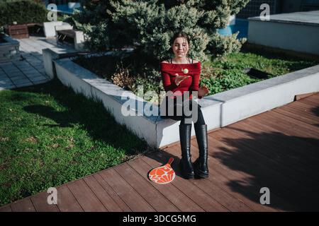 Giovane donna seduta su una sporgenza in cemento all'aperto, con un top rosso a spalla e stivali alti, momenti di gioco Foto Stock