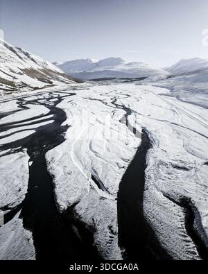 La vista aerea dei fiumi neri si snoda attraverso l'incontaminato paesaggio bianco innevato, annidato tra aspre montagne sotto un cielo limpido, Vik, Wes Foto Stock
