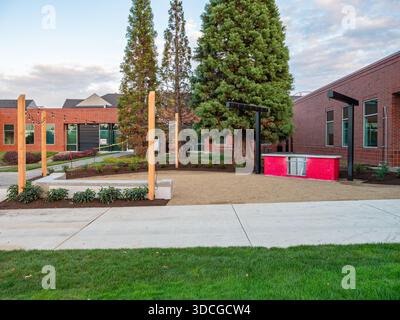 Vista delle panchine rosse che si aprono contro i toni tenui degli edifici in mattoni e degli alberi torreggianti sotto un cielo morbido e nuvoloso ad Apollo Park, Oregon, Stati Uniti. Foto Stock