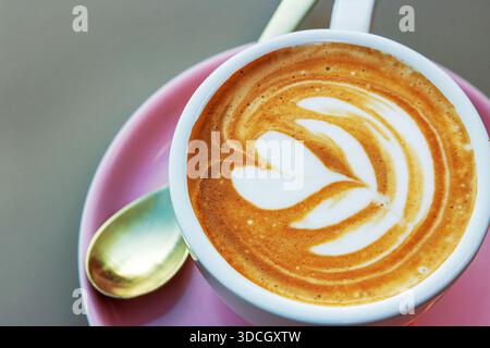 Vista dall'alto di un cappuccino ricoperto di latte art a forma di cuore e fiore in una tazza bianca su un piattino rosa con un cucchiaio accanto. Focu morbida Foto Stock