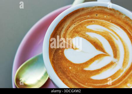 Vista dall'alto di un cappuccino ricoperto di latte art a forma di cuore e fiore in una tazza bianca su un piattino rosa con un cucchiaio accanto. Focu morbida Foto Stock