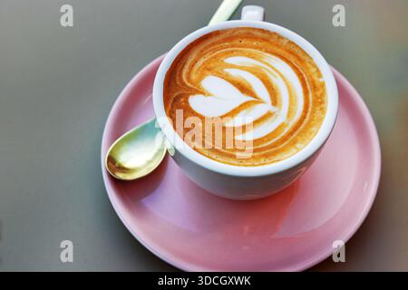 Una tazza di cappuccino con latte art a forma di fiore di cuore su un piattino rosa con un cucchiaio piccolo. Vista dall'alto su una superficie del tavolo liscia. Foto Stock