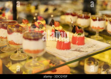 Diversi tipi di dessert sono disposti ordinatamente su un ripiano di vetro in una panetteria. Le persone passano e guardano le gustose prelibatezze, godendo dell'atmosfera di Foto Stock