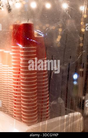Tazze da caffè in carta rossa e bianca impilate dietro una pellicola di plastica bagnata. Luci bokeh calde e sfocate sullo sfondo che aggiungono un'atmosfera accogliente, moderna e autentica Foto Stock