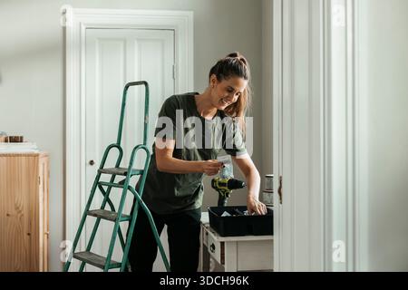 Donna sorridente che prepara la macchina perforatrice stando vicino alla scala in una nuova casa Foto Stock