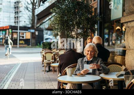 Donna anziana sorridente che indossa cuffie intrauricolari wireless utilizzando un tablet PC mentre è seduta in un bar sul marciapiede Foto Stock
