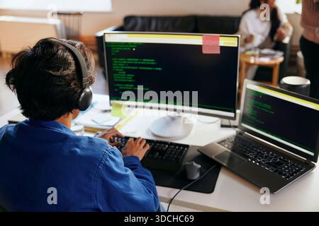 Vista posteriore del giovane programmatore maschio che indossa la codifica delle cuffie wireless sul PC desktop in ufficio Foto Stock
