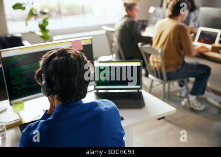 Vista posteriore di un giovane programmatore maschio che indossa la codifica delle cuffie wireless sul computer mentre è seduto alla scrivania in ufficio Foto Stock