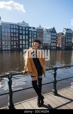 Una donna elegante indossa un abito alla moda di fronte alle iconiche case danzanti Damrak di Amsterdam, Paesi Bassi. Lei è allegra viaggiatrice. Foto Stock