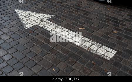 Una freccia bianca dipinta su una strada acciottolata che indica la direzione. La superficie mostra segni di usura e cadute di foglie. Foto Stock