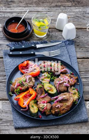 Collo di maiale alla griglia con zucchine arrosto, peperoni e cipolla rossa sul piatto. Servito con salsa di pomodoro, acqua con limone e posate su legno rustico Foto Stock