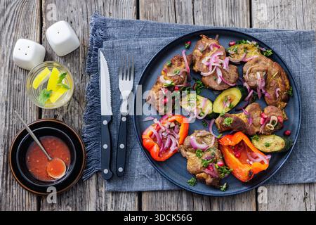 Collo di maiale alla griglia con zucchine arrosto, peperoni e cipolla rossa sul piatto. Servito con salsa di pomodoro, acqua con limone e posate su legno rustico Foto Stock