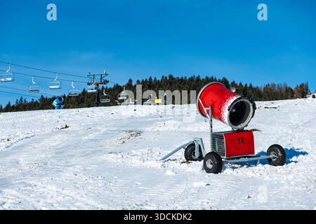 Il cannone da neve rosso si erge su una pista da sci soleggiata a Zakopane, Polonia, garantendo condizioni ottimali per gli sport invernali. Le seggiovie vuote salgono per Foto Stock
