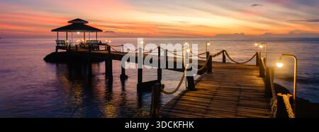 Scoprite il fascino tranquillo dell'isola di Koh Kood in Thailandia mentre il sole tramonta sulle calme acque oceaniche. Un molo in legno con lampade illumina la tranquilla scena, invitante relax. Foto Stock