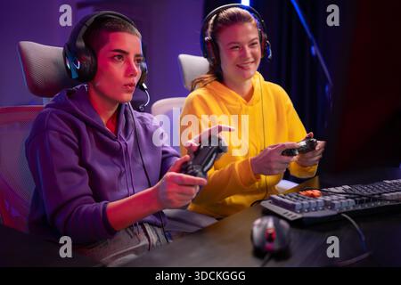Due donne che indossano le cuffie che giocano insieme a un videogioco in una sala giochi colorata. Sono in possesso di controller di gioco, interagiscono con lo schermo e si divertono in una sessione multigiocatore entusiasmante || Model rilasciato Foto Stock