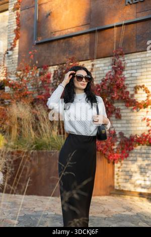 Elegante signora con capelli ricci e occhiali da sole in piedi all'aperto in autunno, vestita con un'accogliente moda stagionale. Foto Stock