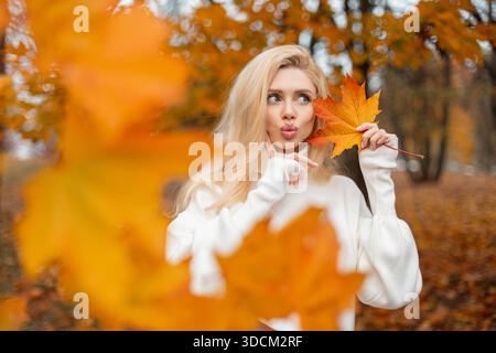 Il brillante ritratto autunnale di una bella ragazza bionda sorpresa con occhi blu e pelle pulita in un maglione bianco alla moda racchiude un te autunnale rosso Foto Stock