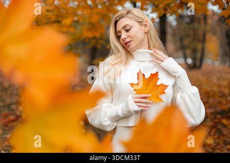 Donna allegra che si gode il giorno d'autunno, tenendo una foglia e in piedi tra alberi dorati. Una ragazza di tendenza positiva ha una brillante foglia di acero giallo colorato. U.S. Foto Stock