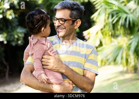 Felice nonno indiano sorridente alla sua nipotina mentre la tiene in un parco verde. Un ritratto caldo e sincero che mette in risalto amore, cura ed emotio Foto Stock