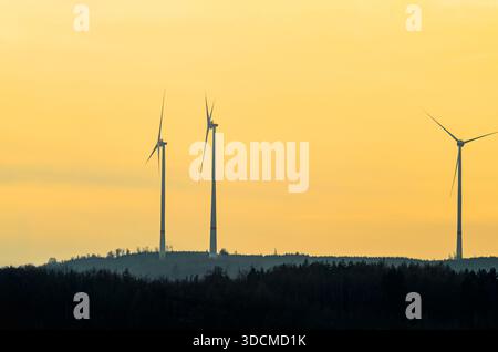 Tre turbine eoliche sono sagomate contro un cielo giallo brillante al crepuscolo, con una linea scura della foresta sotto di esse Foto Stock