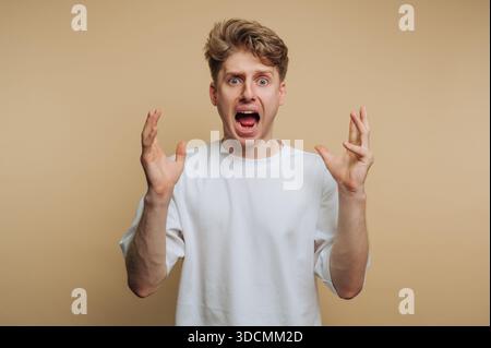Un giovane si trova in uno studio con un background semplice. Ha i capelli marrone chiaro e urla con le mani alzate. La sua espressione mostra una sorpresa Foto Stock