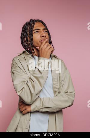 Un giovane si trova in uno studio con uno sfondo rosa. Ha i capelli intrecciati e guarda con attenzione mentre riposa la mano sul mento. Indossa un Foto Stock