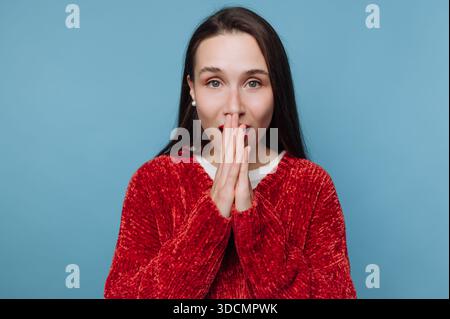 Una giovane donna con lunghi capelli scuri mostra sorpresa mettendo le mani sulla bocca. Si trova su un solido sfondo blu. L'illuminazione è briciola Foto Stock