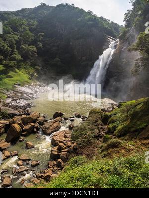 Una vista verticale, ad alto angolo, di una grande e potente cascata che si tuffa da una scogliera scoscesa, scura e rocciosa in un'ampia e turbolenta piscina sottostante, circondata Foto Stock