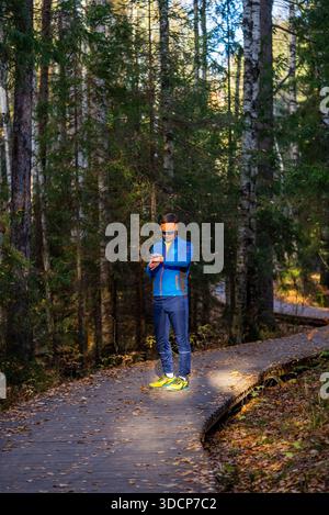 Uomo che fa una pausa su un sentiero del parco, controlla il suo smartwatch fitness per tenere traccia dei progressi dell'allenamento, circondato da colorate foglie autunnali e alberi alti durante un Foto Stock