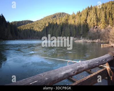 Lago Rosso Romania, lago di montagna ghiacciato circondato da una fitta foresta di pini sotto un cielo limpido, visto da un molo di legno in un tranquillo paesaggio invernale. Foto Stock