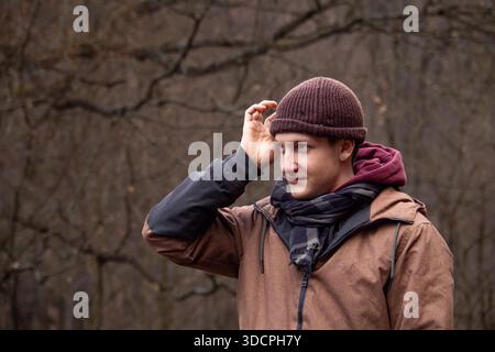 Giovane uomo che indossa un berretto e una giacca in piedi all'aperto in una foresta, adattando il suo cappello durante una fredda giornata autunnale Foto Stock