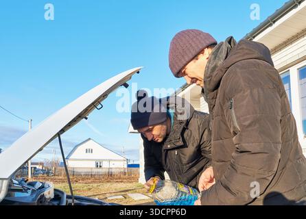 Vista dall'angolazione ridotta dell'anziano e del figlio adulto, vestiti con giacche e tappi caldi, versando liquido lavavetri dalla bottiglia di plastica nel motore dell'auto Foto Stock