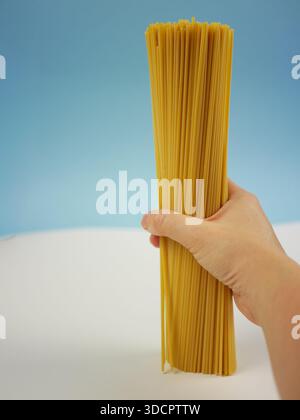 Questo set fotografico mostra gli spaghetti crudi su sfondo blu e bianco. Alcune immagini mostrano una mano che regge un grande fascio di pasta lunga secca Foto Stock