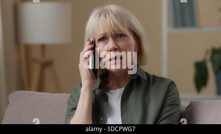 Confuso triste donna caucasica anziana che parla telefono cellulare conversazione negativa a casa sul divano signora in pensione nonna senior female talk Foto Stock