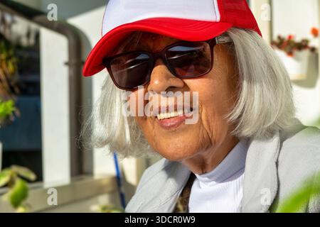 Ritratto ravvicinato di una donna anziana sorridente con capelli bianchi che indossano un berretto rosso e occhiali da sole all'aperto alla luce del sole. L'immagine rappresenta un'immagine attiva Foto Stock