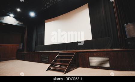 Palcoscenico vuoto con grande schermo di proiezione vuoto, tende nere, gradini in legno, plafoniera e altoparlante audio, pronto per l'uso Foto Stock